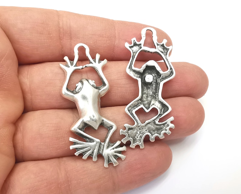 Pode incluir: Dois pingentes de sapo de prata com detalhes elaborados. Os sapos est&atilde;o em posi&ccedil;&atilde;o elevada com as pernas esticadas. Os pingentes t&ecirc;m um la&ccedil;o na parte superior para prender a joias ou outros artesanatos.