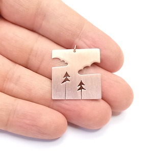 Puede incluir: Un colgante de plata con un diseño recortado de una escena de bosque con árboles y nubes. El colgante cuelga de una cadena de plata.