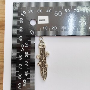 Op de afbeelding: Een zilverkleurige bloemenhanger, ongeveer 5 cm lang, met een gedetailleerd bloemen- en bladerontwerp. De hanger wordt naast een liniaal getoond voor de schaal, met metingen in millimeters.