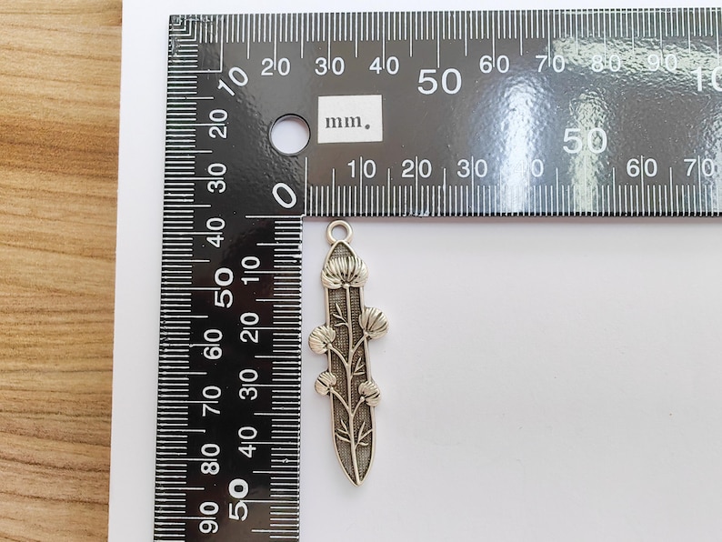 Op de afbeelding: Een zilverkleurige bloemenhanger, ongeveer 5 cm lang, met een gedetailleerd bloemen- en bladerontwerp. De hanger wordt naast een liniaal getoond voor de schaal, met metingen in millimeters.