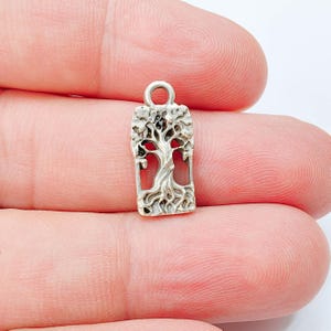Peut inclure: Un petit pendentif rectangulaire en métal argenté présentant un motif en relief détaillé représentant un arbre avec des racines et des branches visibles. Le pendentif possède une boucle circulaire sur le dessus pour la fixation.