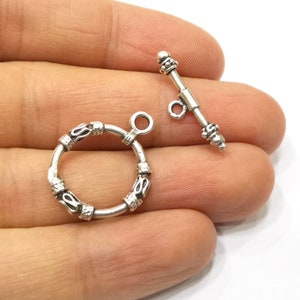 Könnte beinhalten: Zwei silberfarbene Knebelverschlüsse. Einer ist ein kreisförmiger Ring mit dekorativen Details, der andere ein gerader Stab mit einer Schlaufe und dekorativen Enden. Diese Verschlüsse sind wahrscheinlich für die Schmuckherstellung.