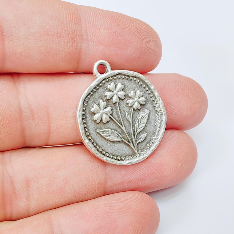 May include: A silver-colored round charm with a floral design. The charm features three flowers with detailed petals and leaves, set within a beaded border. It has a small loop at the top for attaching to a chain or bracelet.