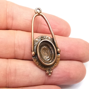 May include: A copper-toned metal pendant with an oval-shaped opening. The pendant has a decorative design with a raised edge and a textured surface.