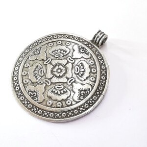 May include: A silver-colored, round pendant with an intricate floral design. The pendant features a central flower surrounded by stylized petals and a border of small circles. A loop is attached at the top for a chain.