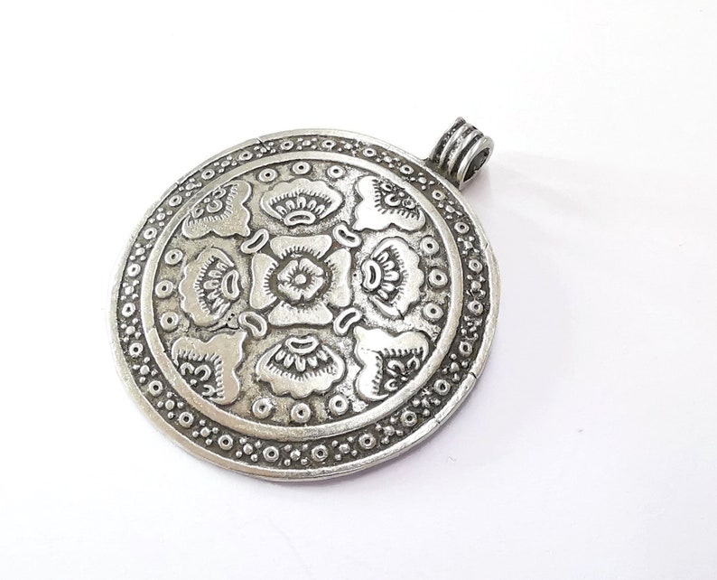 May include: A silver-colored, round pendant with an intricate floral design. The pendant features a central flower surrounded by stylized petals and a border of small circles. A loop is attached at the top for a chain.