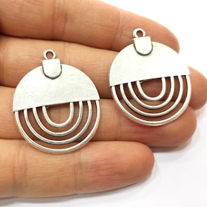 Op de afbeelding: Twee zilverkleurige metalen hangerbedels met een cirkelvormig ontwerp. Elke hanger heeft een halfronde bovenkant met een kleine lus aan de bovenkant en een reeks concentrische cirkels in de onderste helft.