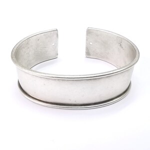 Puede incluir: Una pulsera de puño plateada con un diseño redondeado. La pulsera tiene una banda ancha con un borde decorativo en relieve y un diseño abierto. El metal tiene un acabado cepillado y la pulsera está sobre un fondo blanco.
