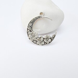 May include: A silver-toned crescent moon pendant with floral detailing. The pendant features a crescent shape with a textured surface and is adorned with several flower designs. It has a loop at the top for hanging.