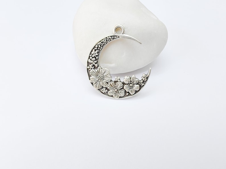 May include: A silver-toned crescent moon pendant with floral detailing. The pendant features a crescent shape with a textured surface and is adorned with several flower designs. It has a loop at the top for hanging.