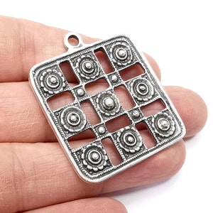 May include: Silver pendant with a square design featuring a geometric pattern of circles and squares. The pendant has a textured surface and a silver finish.