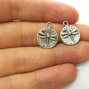 May include: Two silver-toned metal charms in the shape of a circle with a dragonfly design. The dragonfly is raised and detailed.