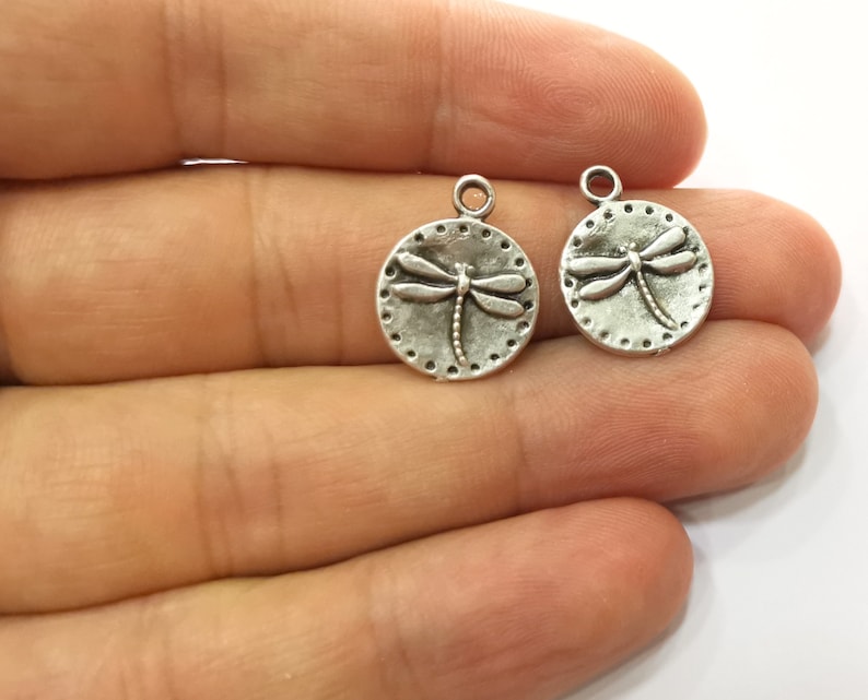 May include: Two silver-toned metal charms in the shape of a circle with a dragonfly design. The dragonfly is raised and detailed.