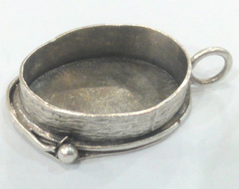 May include: A silver-colored oval pendant setting with a textured rim and a small loop for a chain. The setting has a decorative design element on one side, featuring a small ball and curved lines.