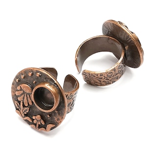 May include: Two copper adjustable rings with a textured finish. The rings have a round bezel setting for a cabochon stone. One ring has a floral design with a moon and stars.