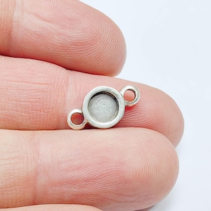 May include: A small, silver-coloured jewellery finding with a circular bezel and two loops. The finding is held between fingers against a white background. The bezel is designed to hold a small stone or other decorative element.