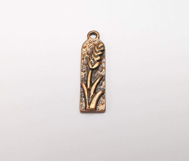 May include: A copper-toned metal charm with a wheat stalk design. The charm is rectangular and has a textured surface.