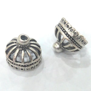 Pode incluir: Duas contas prateadas em forma de coroa. As contas têm um design detalhado com barras verticais e uma faixa decorativa ao redor da base. Uma conta está em pé, e a outra está inclinada, mostrando os detalhes intrincados.