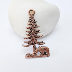 May include: A copper charm featuring a detailed pine tree with a bear standing beneath it. The charm has a loop at the top for hanging.