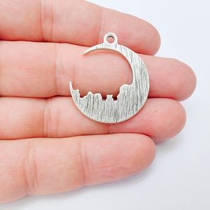 May include: Silver-toned crescent moon pendant with a textured surface and a small loop for hanging. The moon shape features a silhouette of three figures along the bottom edge. Ideal for jewelry making or crafting projects.