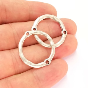 May include: Two silver-colored, hammered-look metal circle connectors with three holes each. The connectors are held in a hand against a white background. These jewelry findings are ideal for crafting earrings, necklaces, or other accessories.