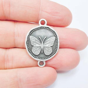 Könnte beinhalten: Ein silberfarbener Metallanhänger mit einem Schmetterlingsdesign. Der runde Anhänger hat einen Perlenrand und zwei Schlaufen zur Befestigung. Der Schmetterling ist mittig platziert, mit detaillierten Flügelmustern.