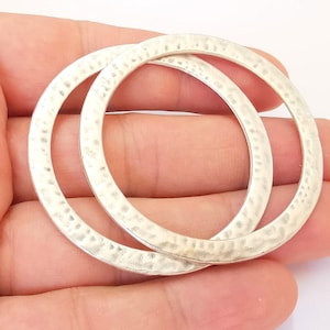 May include: Two silver hammered metal hoops, one slightly larger than the other, are shown in a hand. The hoops are smooth and have a textured surface.