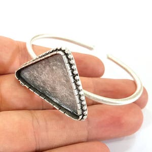 Può includere: Un braccialetto argentato con un'incastonatura triangolare. L'incastonatura ha una superficie strutturata e chiara, bordata da piccole perle. Il braccialetto ha un design a polsino aperto.