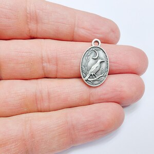May include: Silver charm featuring a detailed design of a crow perched on a branch with a crescent moon in the background.