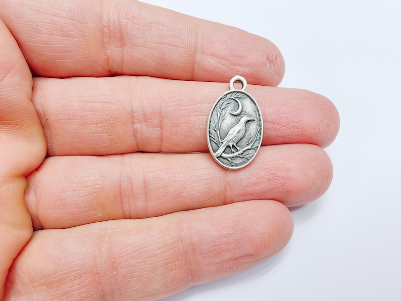 May include: Silver charm featuring a detailed design of a crow perched on a branch with a crescent moon in the background.