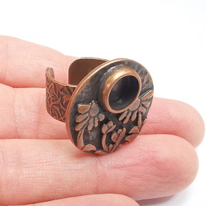 May include: A copper ring with a floral design and a round opening in the center. The ring is worn on a finger.