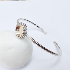 May include: A silver-toned cuff bracelet with a circular setting. The setting is encrusted with small, clear crystals around a gold-colored interior. The bracelet is resting on a white, rounded stone.