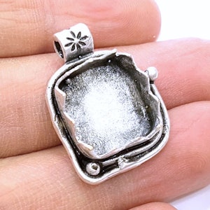 May include: Silver pendant setting with a square bezel and a textured border. The setting has a bail with a flower design and two small balls on the sides.