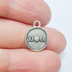 Puede incluir: Un pequeño colgante plateado con un diseño de fases lunares. El colgante tiene forma circular con un borde de cuentas y un lazo en la parte superior para sujetarlo a una cadena o cordón. Las fases lunares están representadas en el centro.