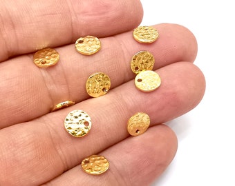 May include: A collection of small, hammered gold-colored metal disc beads. Each disc has a textured surface and a small hole for stringing. The beads are scattered across a light background.