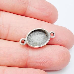 Op de afbeelding: Ovale zilverkleurige bezel setting met twee lussen aan weerszijden. De bezel heeft een ondiep, verzonken gebied voor het plaatsen van een steen of ander versiering. De setting wordt in een hand gehouden.