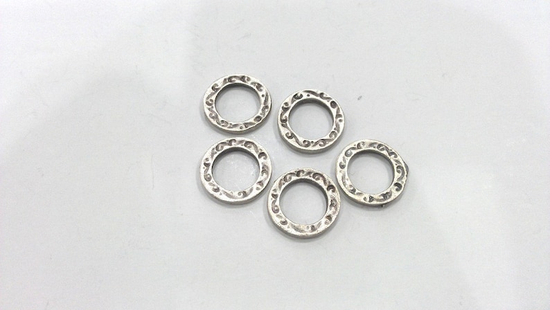 May include: Five silver jump rings with a decorative, ornate design. The rings are all the same size and are arranged in a loose circle.