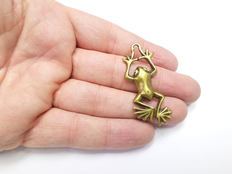 May include: A gold-toned metal frog charm with a loop at the top for attaching to a necklace or bracelet. The frog is in a leaping pose with its legs outstretched.