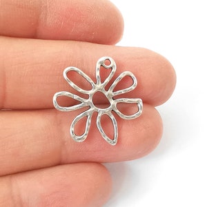 Puede incluir: Un charm de flor calada de color plateado con textura martillada. El charm tiene un pequeño lazo en la parte superior para sujetarlo a la joyería. La flor tiene ocho pétalos y un centro circular.