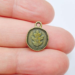May include: A small, antique brass-colored charm with an oak leaf design. The charm is round with a beaded border and a loop at the top for attaching to a necklace or bracelet. The oak leaf is in the center of the charm.
