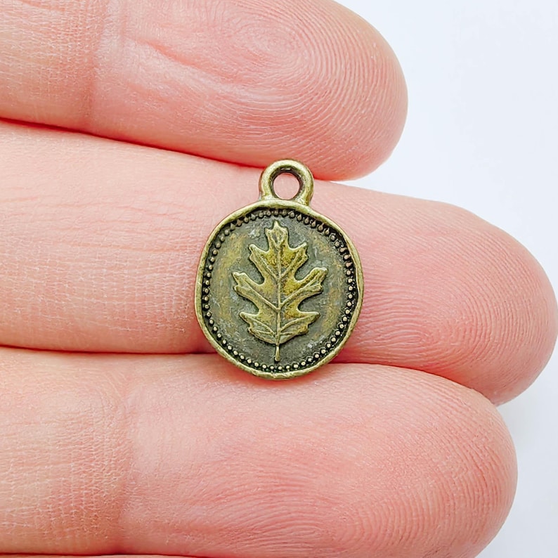 May include: A small, antique brass-colored charm with an oak leaf design. The charm is round with a beaded border and a loop at the top for attaching to a necklace or bracelet. The oak leaf is in the center of the charm.