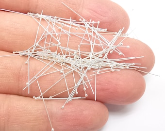 May include: A collection of silver-colored head pins with small, round heads. The pins are scattered across a light-colored surface, with many of them appearing to be held in a hand. The pins are straight and have a shiny finish.
