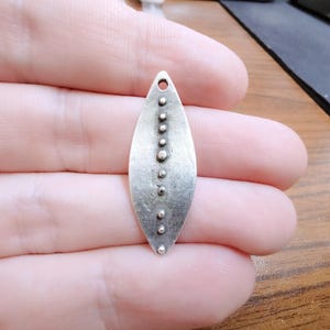 Puede incluir: Un colgante de plata con forma de hoja con una fila de pequeños puntos de plata en el centro.