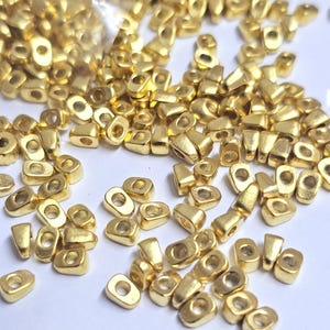 May include: A collection of small, gold-colored metal beads. Each bead has a rectangular shape with a rounded top and a hole running through the center. The beads are scattered across a white surface, some spilling from a clear plastic bag.