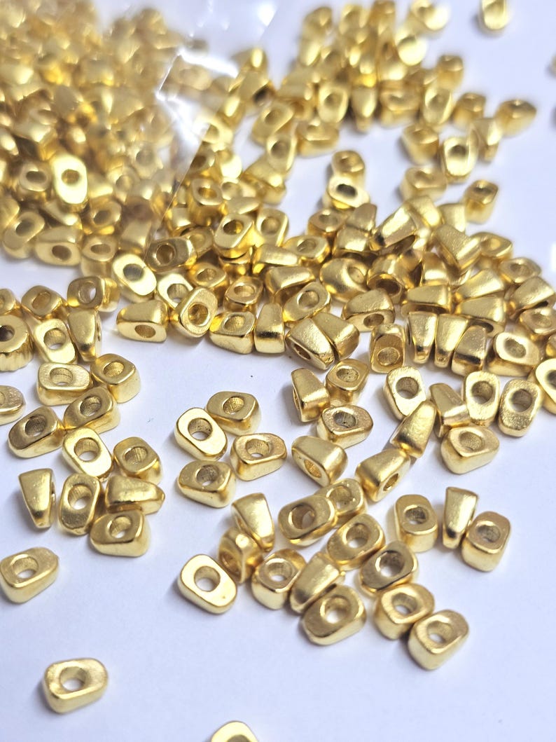 May include: A collection of small, gold-colored metal beads. Each bead has a rectangular shape with a rounded top and a hole running through the center. The beads are scattered across a white surface, some spilling from a clear plastic bag.