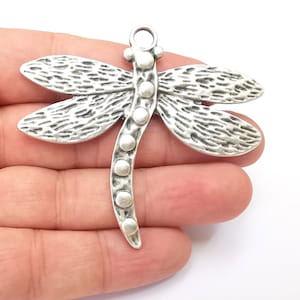 Op de afbeelding: Zilverkleurige libellenbedel met een getextureerd lichaam en een kralenstaart. De bedel heeft een lus aan de bovenkant om aan sieraden of andere knutselwerkjes te bevestigen.