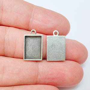 May include: Two rectangular silver-toned pendant blanks with a loop at the top for attaching to a chain or cord. One blank has a recessed area for setting a stone or other element. The other is a solid rectangle.