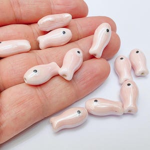 Puede incluir: Una colección de pequeñas cuentas de cerámica rosa con forma de pez y ojos de punto negro. Las cuentas son lisas y brillantes, con un agujero para ensartar. Los peces están en varias posiciones. Ideal para la fabricación de joyas.
