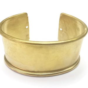 Op de afbeelding: Een brede, open manchetarmband in een geborstelde goudtint. De manchet heeft een afgeronde boven- en onderrand, met een platte centrale band. De armband is gemaakt van metaal en heeft een eenvoudig, elegant ontwerp. De armband is ongeveer 5 cm breed.