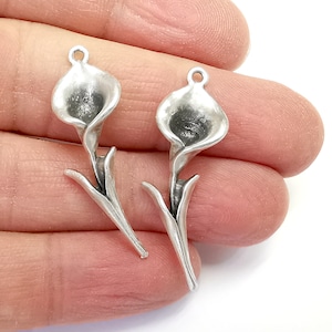 May include: Two silver-toned metal charms shaped like calla lilies. The charms have a detailed design and are perfect for jewelry making or crafting.